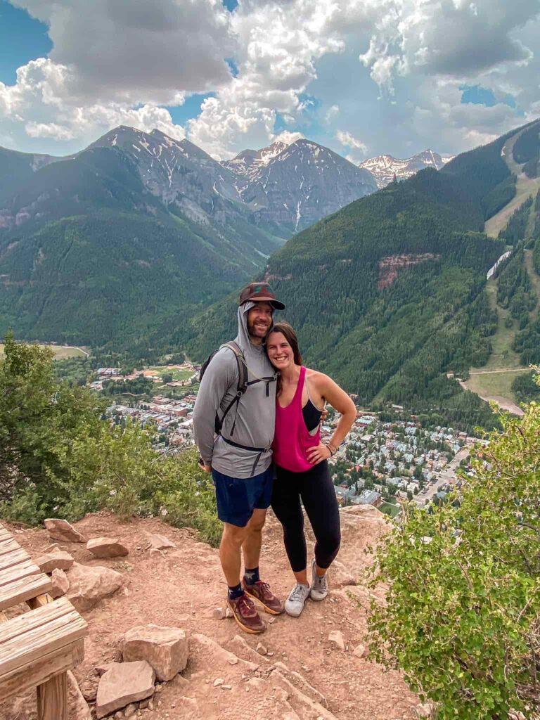 A man and woman stand on a rcok overlooking the town of Telluride with mountains in the distance while hiking the Jud Weibe Memorial Trail.