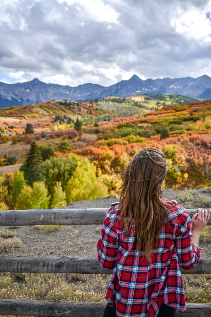 Woman in a red plaid shirt overlooking fall foliage and mountains in the distance while leaf peeping in Colorado.