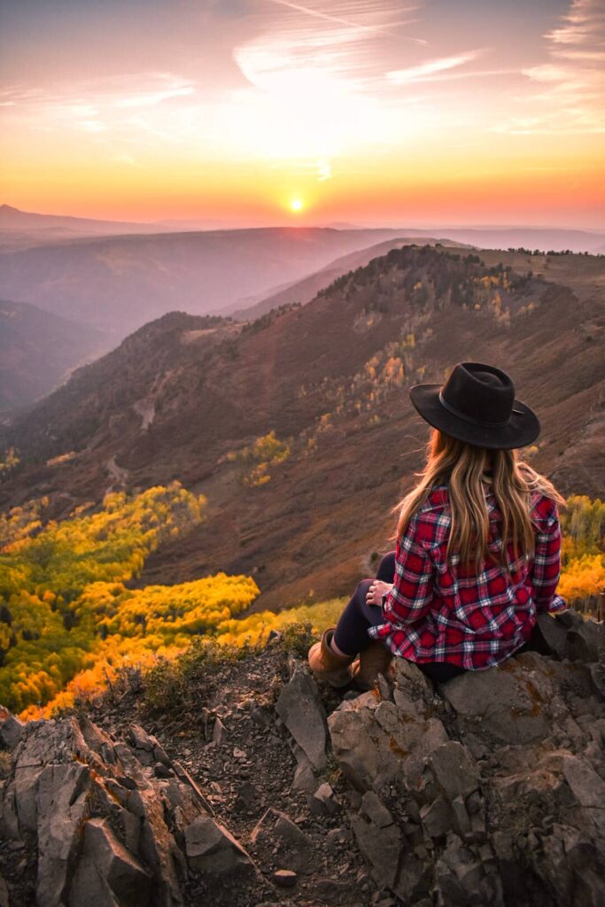 Woman wearing a plaid shirt and wide brim black hat sits on a rock overlooking golden aspen forests in the mountains near Telluride at sunset.