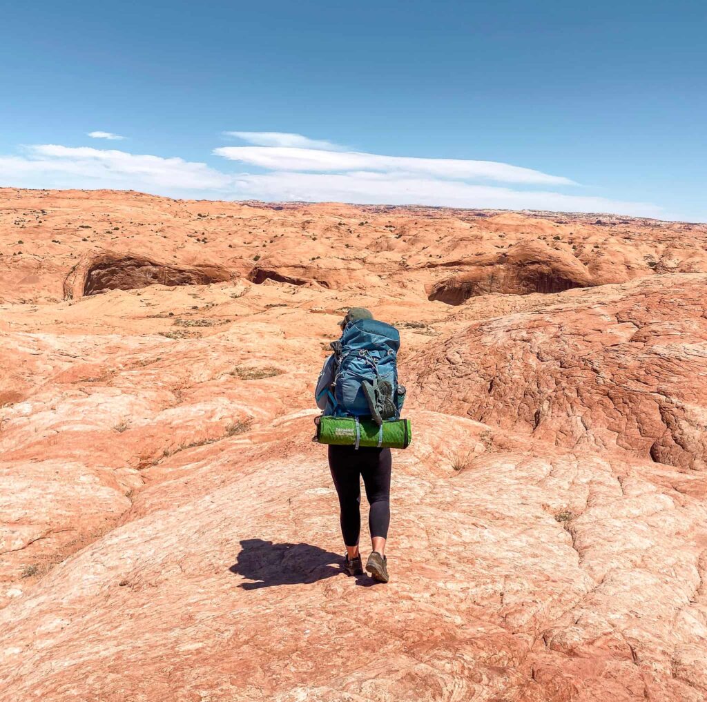 Woman carrying a large blue backpack with a green sleeping pad and sandals attached hikes across orange slickrock in the desert in Escalante, Utah.