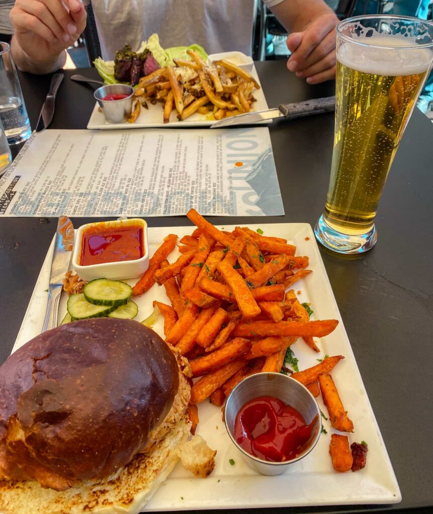 Steak tips and fries, a beer, and a chicken sandwich and sweet potato fries on a table.