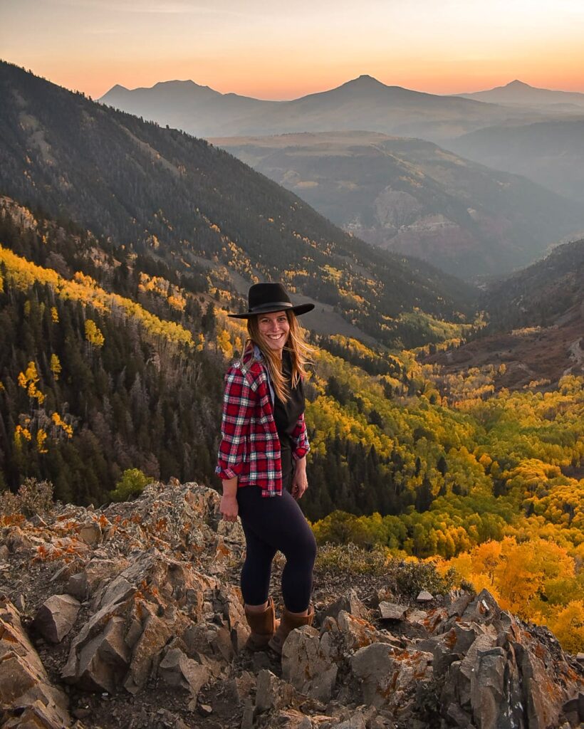 Woman in a red plaid shirt and wide-brim black hat stands on a rock overlooking a golden aspen forest with a hazy sunset over the mountain in the distance in Telluride, Colorado.