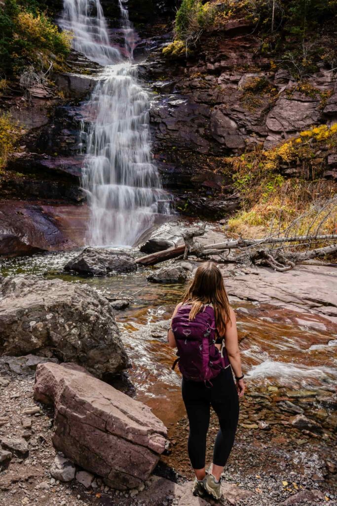 Woman wearing a hiking pack stands in front of a waterfall in Telluride, Colorado.