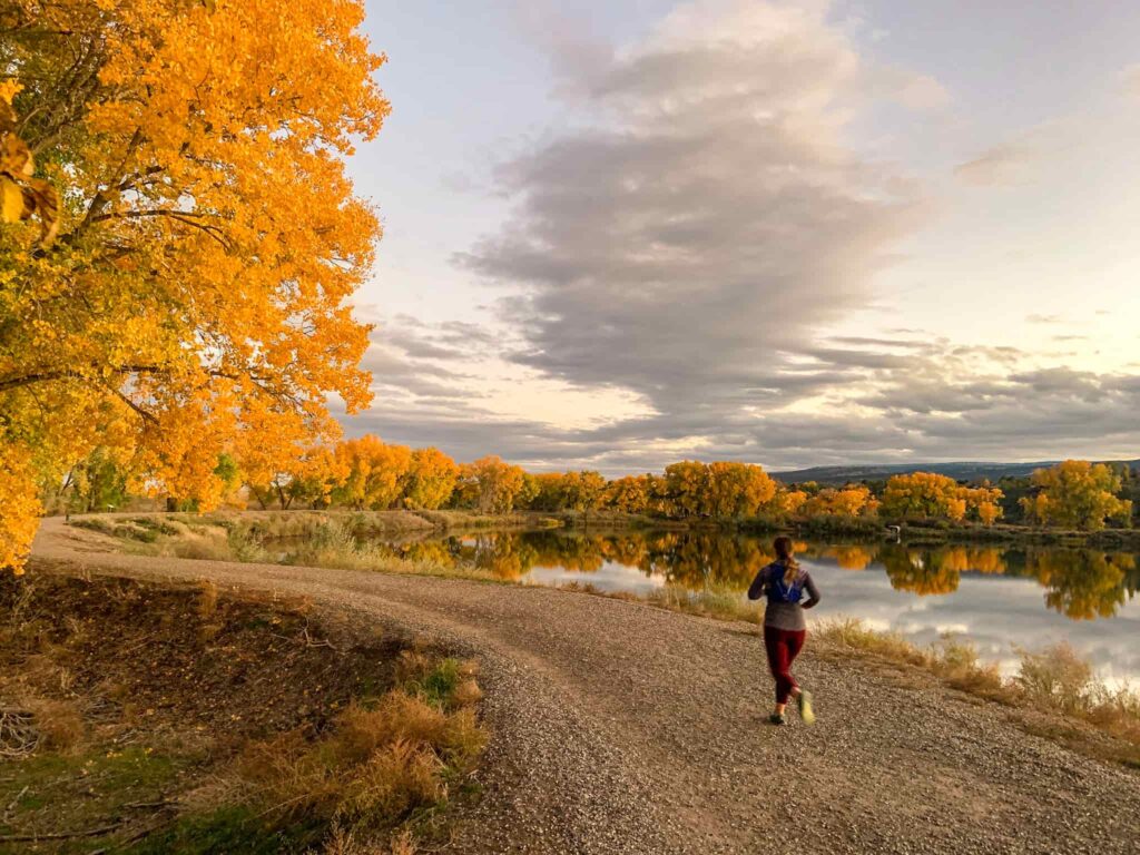 Woman runs on a gravel path next to a lake surrounded by golden cottonwood trees at Connected Lakes in Grand Junction, Colorado.