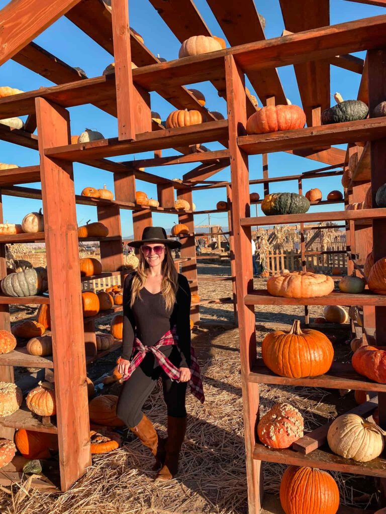 Woman wearing all black with a plaid flannel around her waste and brown knee-high boots stands in a pumpkin house at Studt Farm in Grand Junction.
