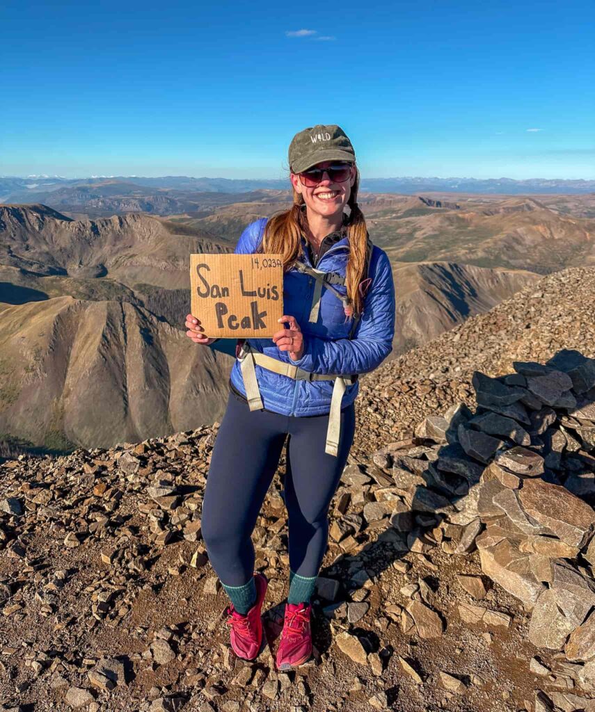 Woman holding a sign that says "San Luis Peak" while standing on top of a mountain with blue skies.