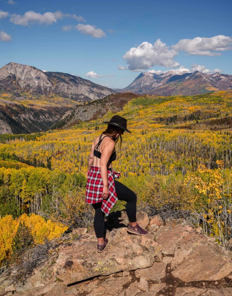 A woman wearing a fall hiking outfit while hiking in Crested Butte, Colorado with golden aspens and mountains behind her.