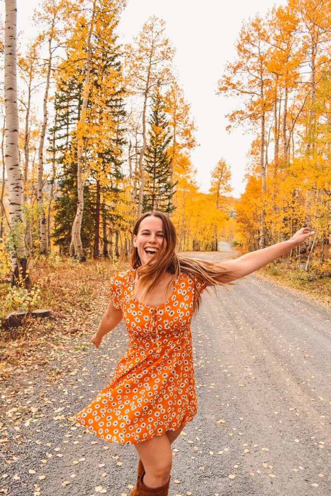 Woman in an orange floral dress dances on a dirt road surrounded by golden aspen trees on the Grand Mesa in the fall.