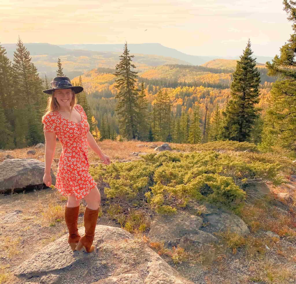 woman in orange floral dress stands on a rock with a golden aspen forest behind her.