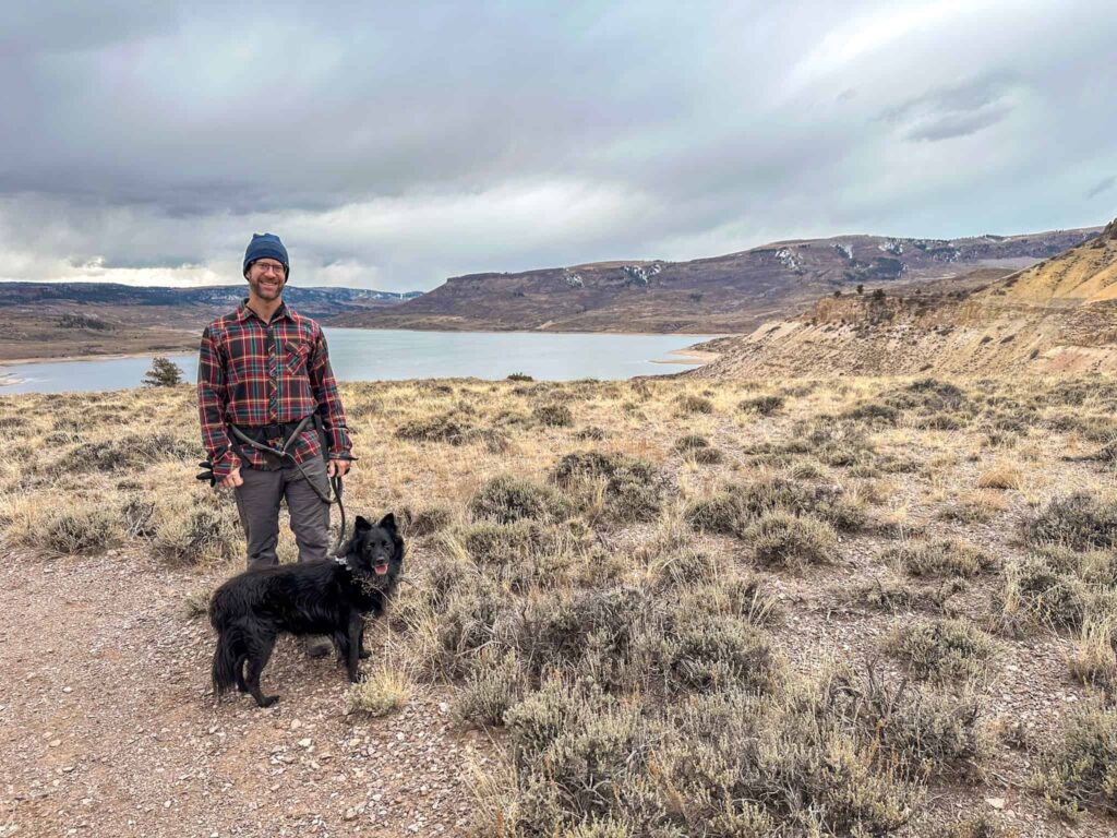 Man hiking with a black dog on a leash in front of a reservoir.