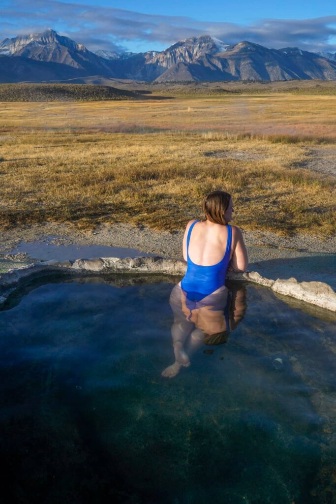 Woman in a blue bathing suit soaking in a hot spring in front of a field with mountains in the distance.