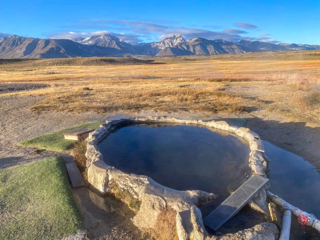 Hilltop Hot Springs near Mammoth Lakes, California. A large pool with a pipe leading to it with snow capped mountains in the distance.