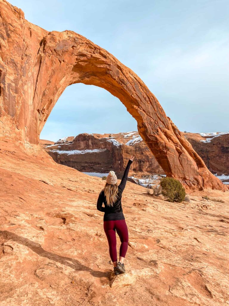 Woman standing with a peace sign in the air in front of Corona arch while hiking in Southern Utah.