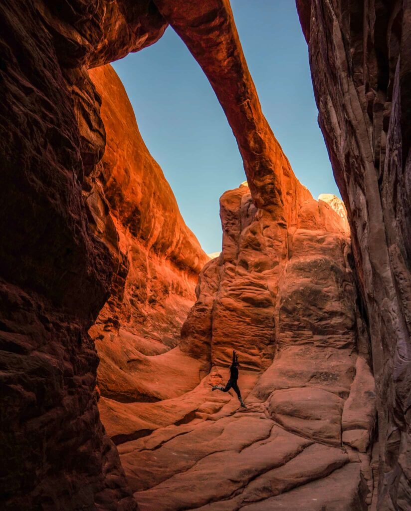 Woman leaping under a sandstone arch while hiking in the Fiery Furnace in Arches National Park.