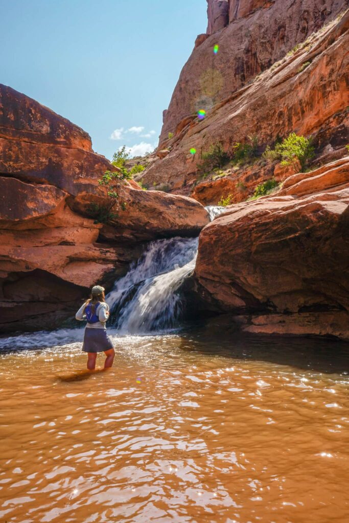 Woman standing in a pool of water in front of a desert waterfall in Moab.