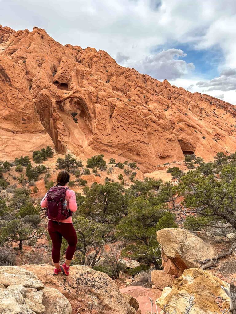 Woman in a purple backpack and hot pink trail runners stands on a rock looking at a wavy sandstone rock formation in Capitol Reef National Park.