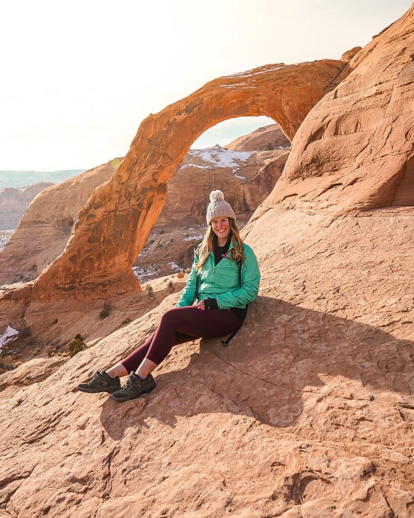Woman dressed for a winter hike sits on slickrock in front of Corona Arch in Moab.
