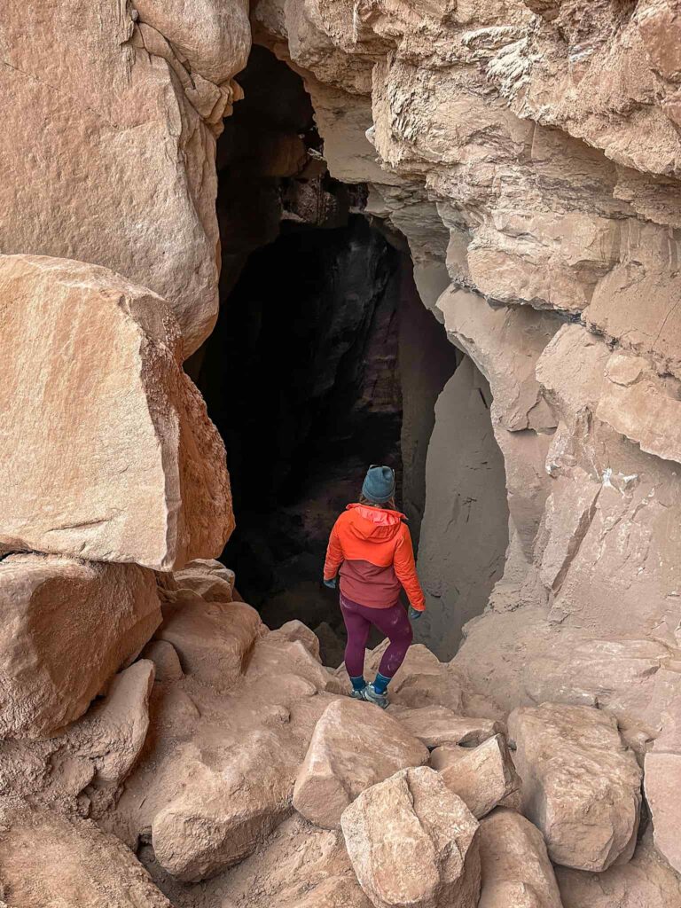Woman in an orange jacket stands in front of a giant cave in Goblin Valley State Park.