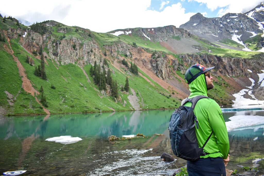 Man in lime green shirt and Colorado hat standing in front of Lower Blue Lake while hiking in Colorado.