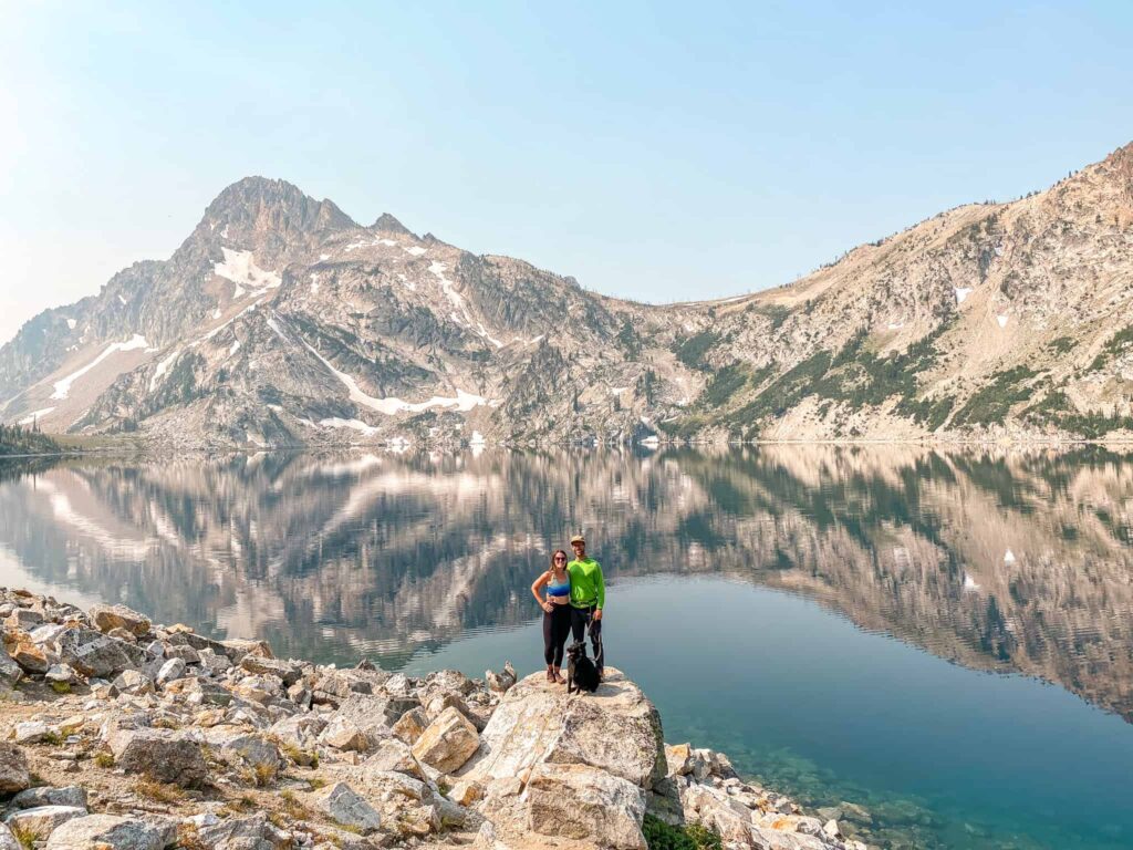 A couple and their dog stand on a rock in front of an alpine lake surrounded by mountains while hiking in Idaho.