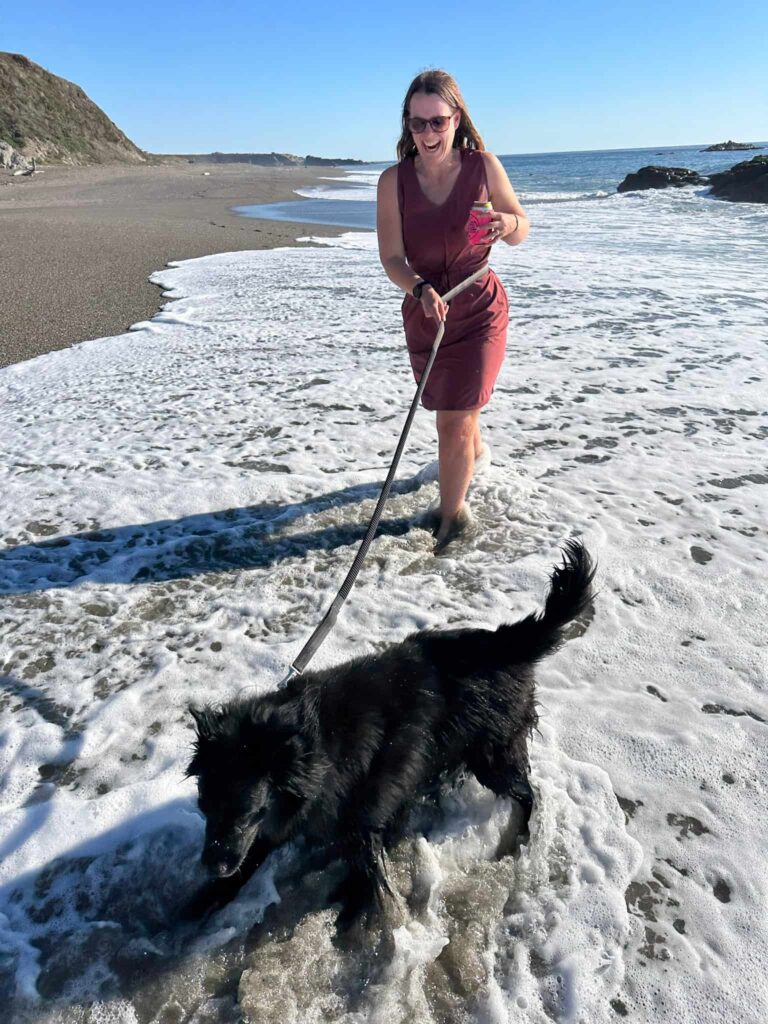 Woman holding a beer and walking through the water at the beach with a leash around her waist walking her black dog.