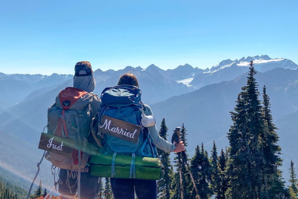 A couple backpacking on their honeymoon in Olympic National Park with mountains in the distance and "just married" signs on their backpacks.