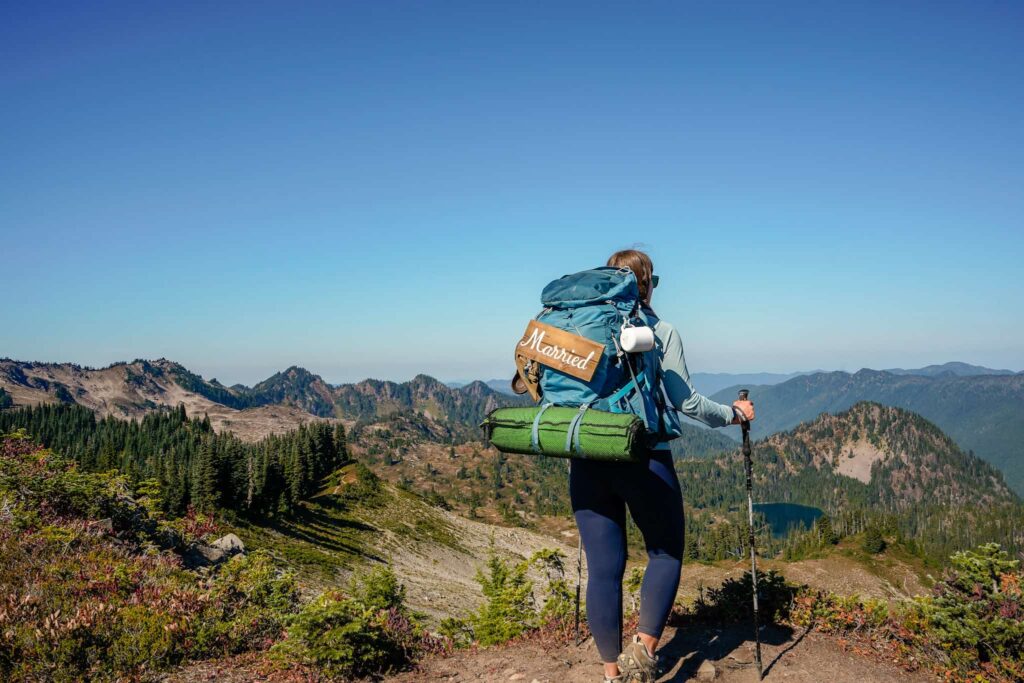 Woman backpacking in Olympic National Park on her honeymoon with a "married" sign on her backpack with mountains and alpine lakes in the distance.