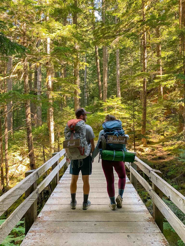 A couple holding hands while crossing a bridge while backpacking in a forest on their honeymoon in Olympic National Park with "just married" signs on their backpacks.