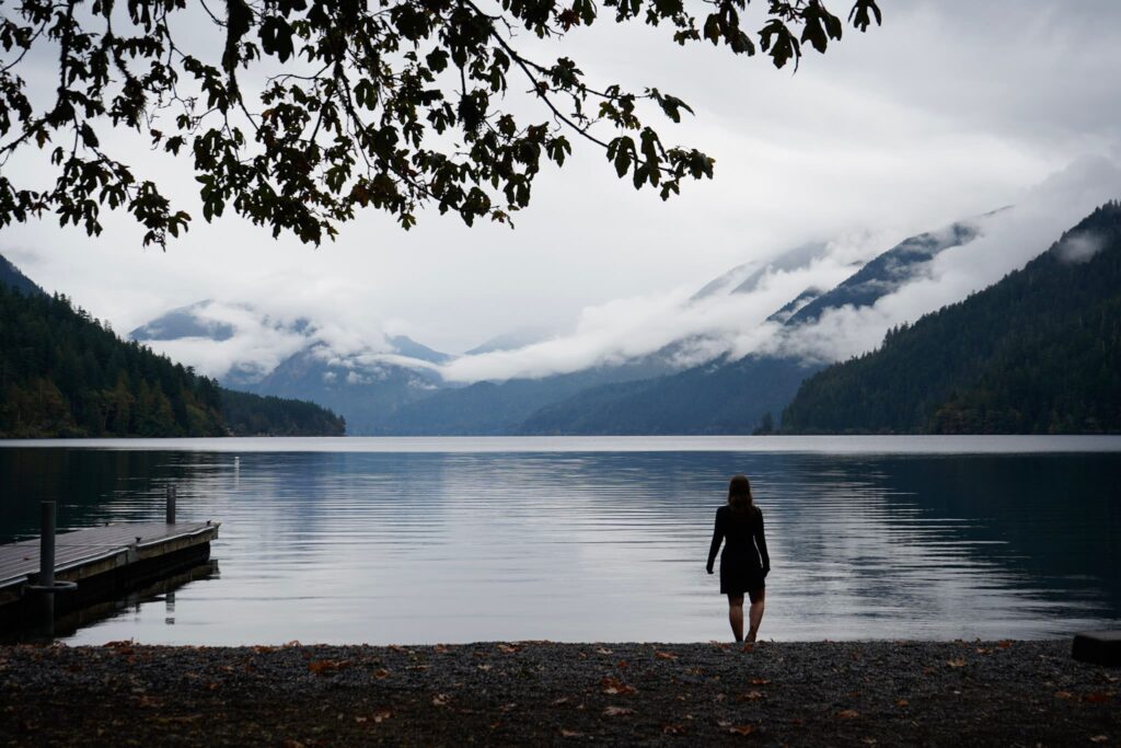 A woman stands on the shore Lake Crescent in Olympic National Park with cloud covered mountains in the distance.