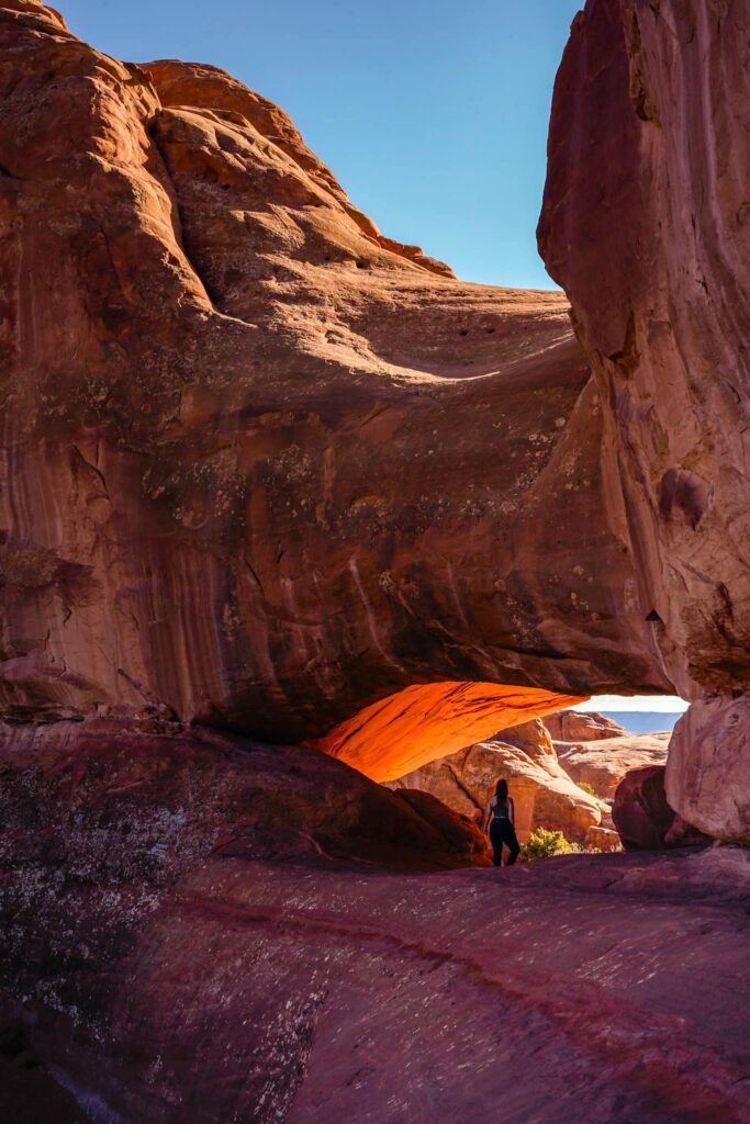 A woman stands in the mouth of a sandstone arch in the desert.