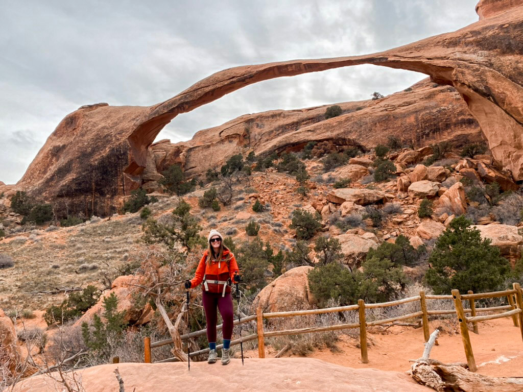 Woman holding hiking poles and dressed for winter stands on a rock in front of a long arch in Arches National Park.