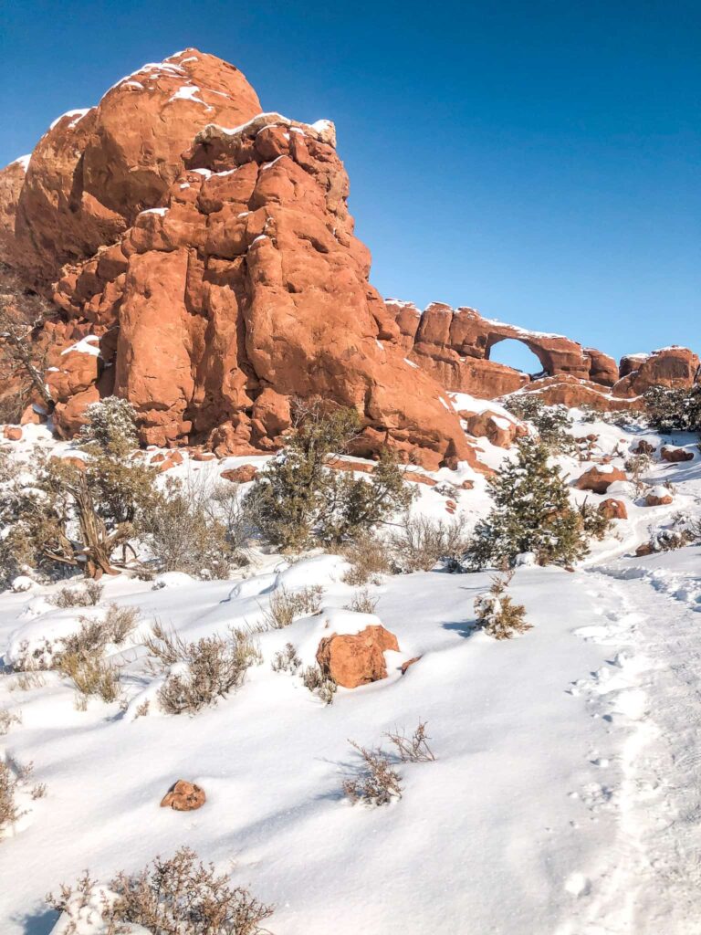 A snowy trail leading to a sandstone arch with sandstone rocks surrounding it.