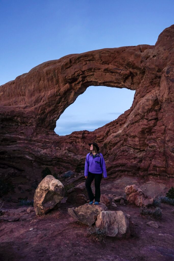 A woman stands on a rock in front of an eye-shaped arch during blue hour.