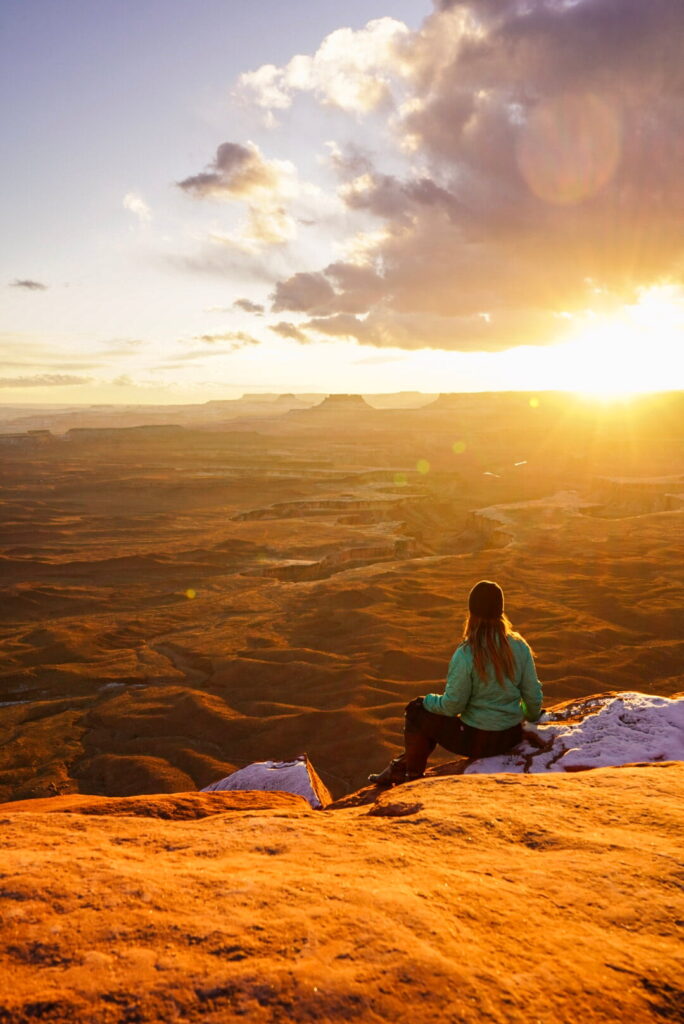 Women sits on the edge of a rock overlooking a network of canyons at sunset in Moab, Utah.