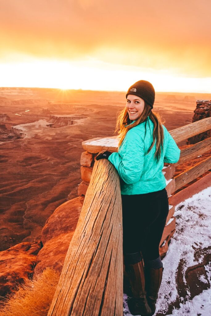 Woman in a mint green jacket and hat looks over her shoulder while leaning on a guardrail overlooking a network of canyons at sunset in Canyonlands National Park.