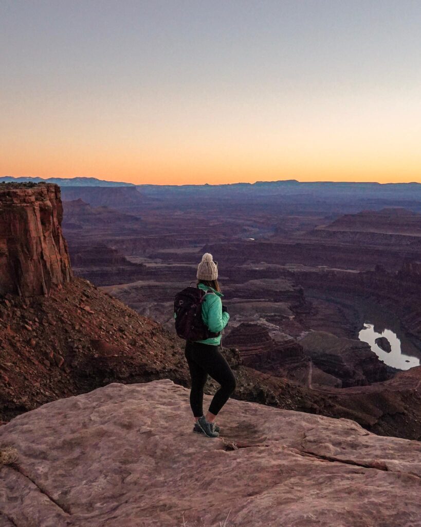 Woman in winter hiking attire stands on a rock overlooking the Colorado River at. the bottom of a canyon at sunset in Moab, Utah.