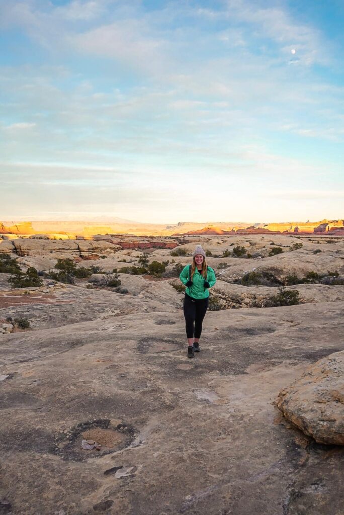 Woman stands on a rock with glowing rocks in the distance at sunset.