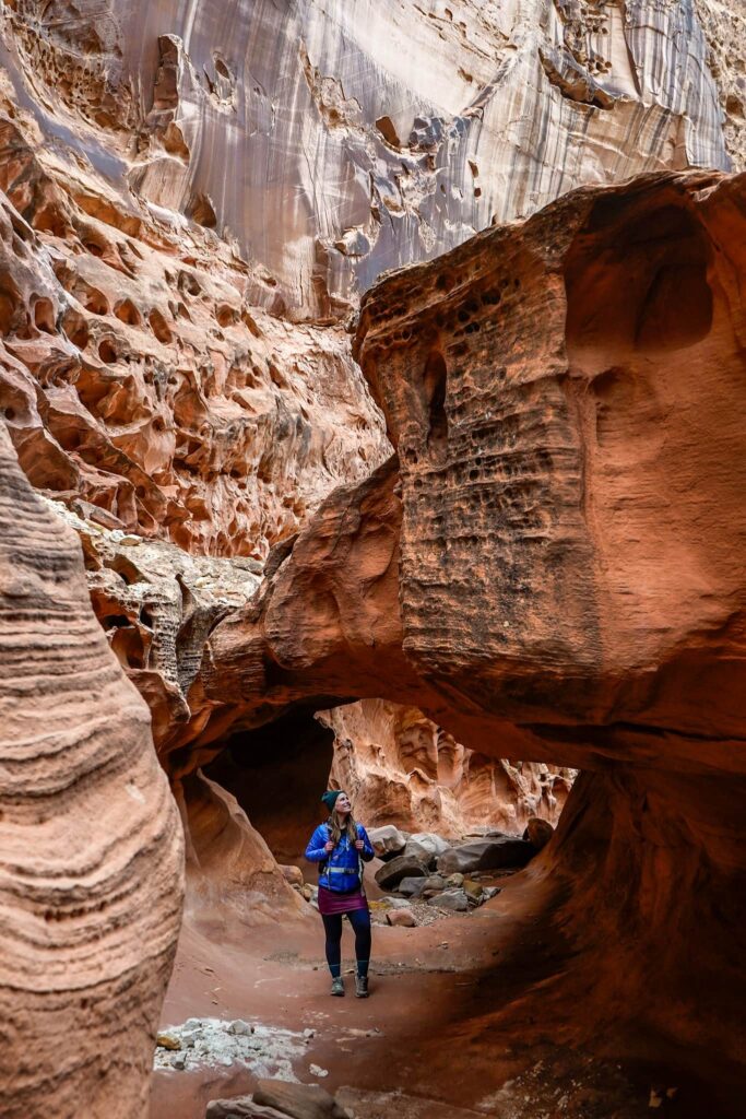 Woman in winter hiking attire stands under a sandstone bridge in a slot canyon in the San Rafael Swell.