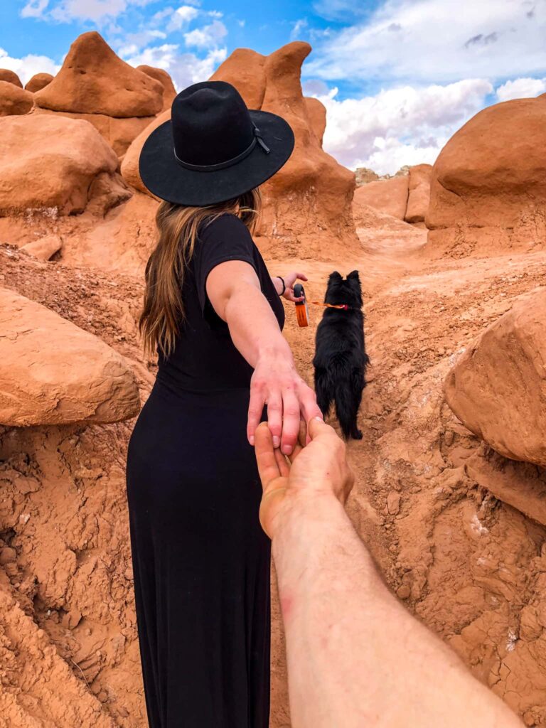 Woman in black dress with black hat reaching back and holding a man's hands while also holding a leash with a black dog while hiking through orange hoodoos in Goblin Valley State Park.