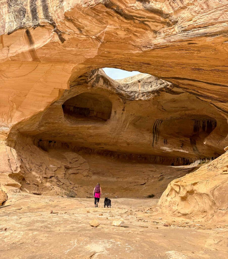 A woman and black dog hike under a large sandstone alcove with a large arch inside it.