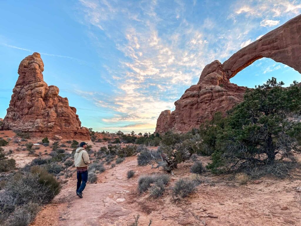 Man stands in the desert with a rock arch to his right and a rock formation behind him.