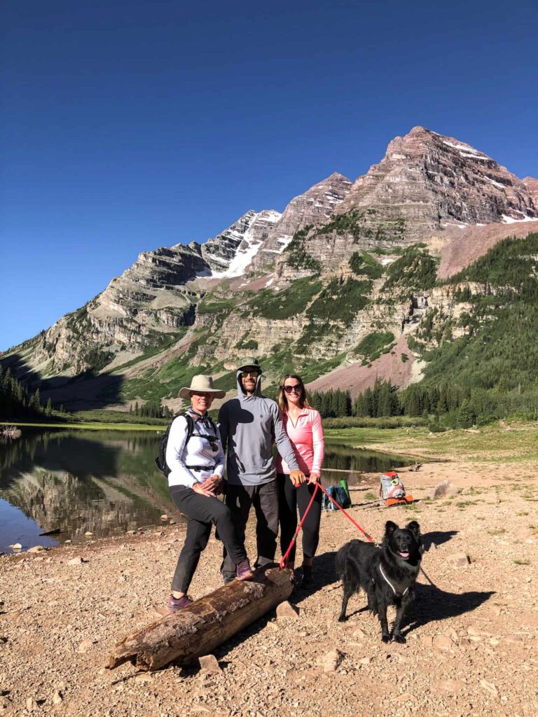 A family and dog stand on the shore of a lake in front of the Maroon Bell mountains in Colorado.