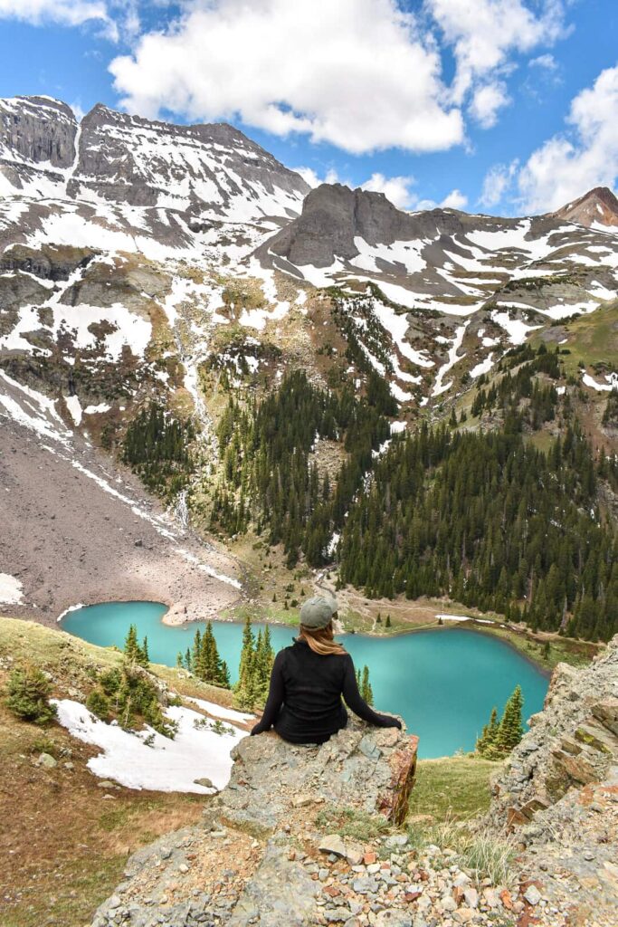 Woman sits on a rock overlooking a turquoise blue lake surrounded by snow-capped mountains in Colorado.