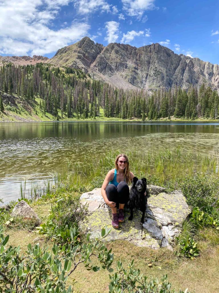 Woman and black dog sitting on a rock in front of a lake with mountains in the background on one of the best dog-friendly hikes in Colorado.