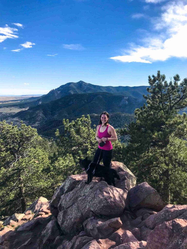 Woman in a pink tank top holding a leash with a black dog standing on a rock in front of mountains in Boulder, Colorado.