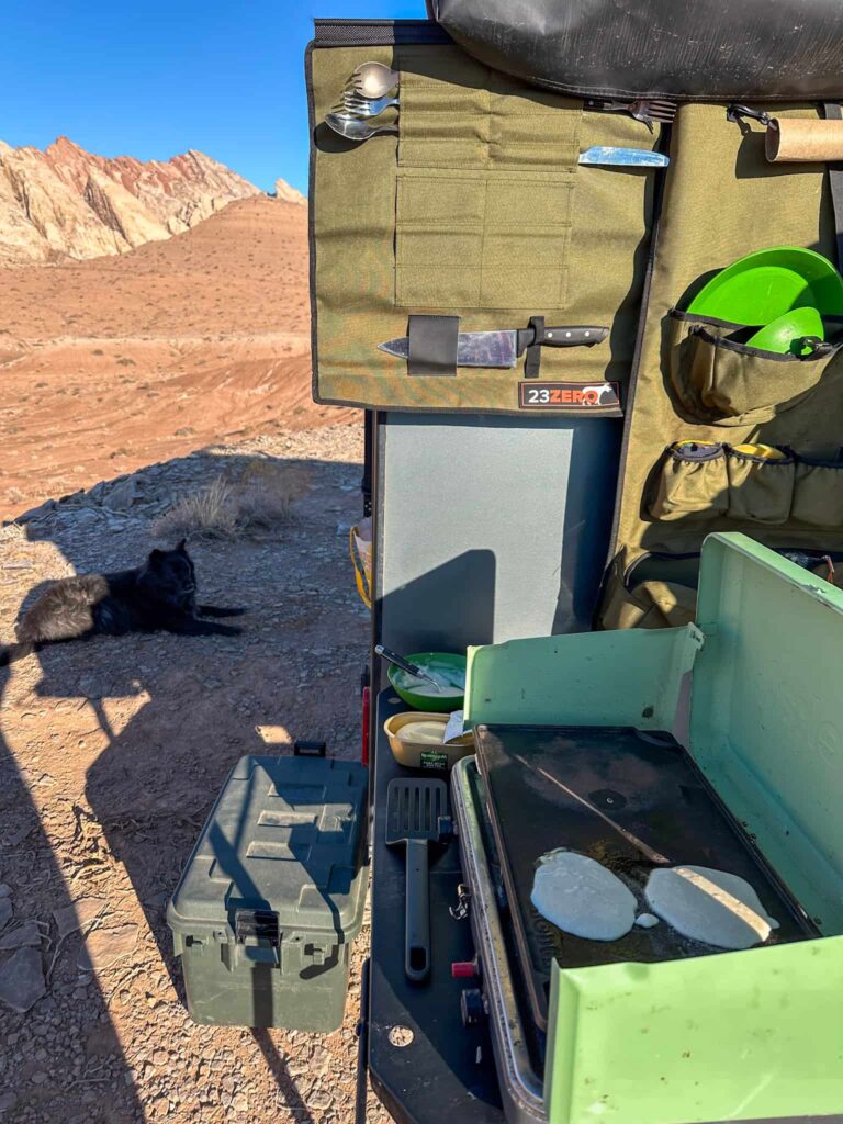 Pancakes cooking on a griddle on top of a green camp stove with a 23Zero kitchen organizer attached to a trailer behind it while in the desert.