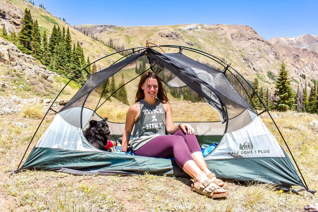 A woman and black dog sitting in an REI half dome 1 plus tent while on a last-minute camping trip in the mountains in Colorado.