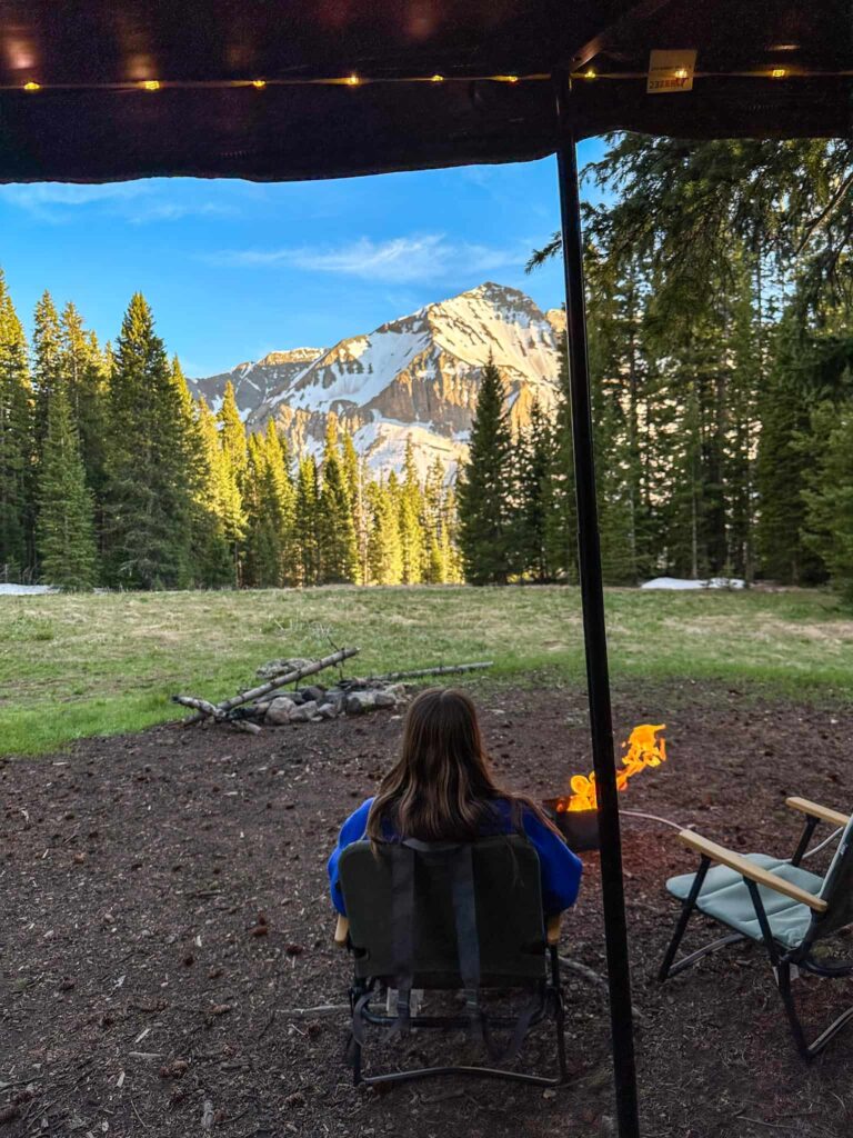 Woman sitting on a camp chair in front of a fire with snow covered mountains in the distance while camping in Telluride, Colorado.