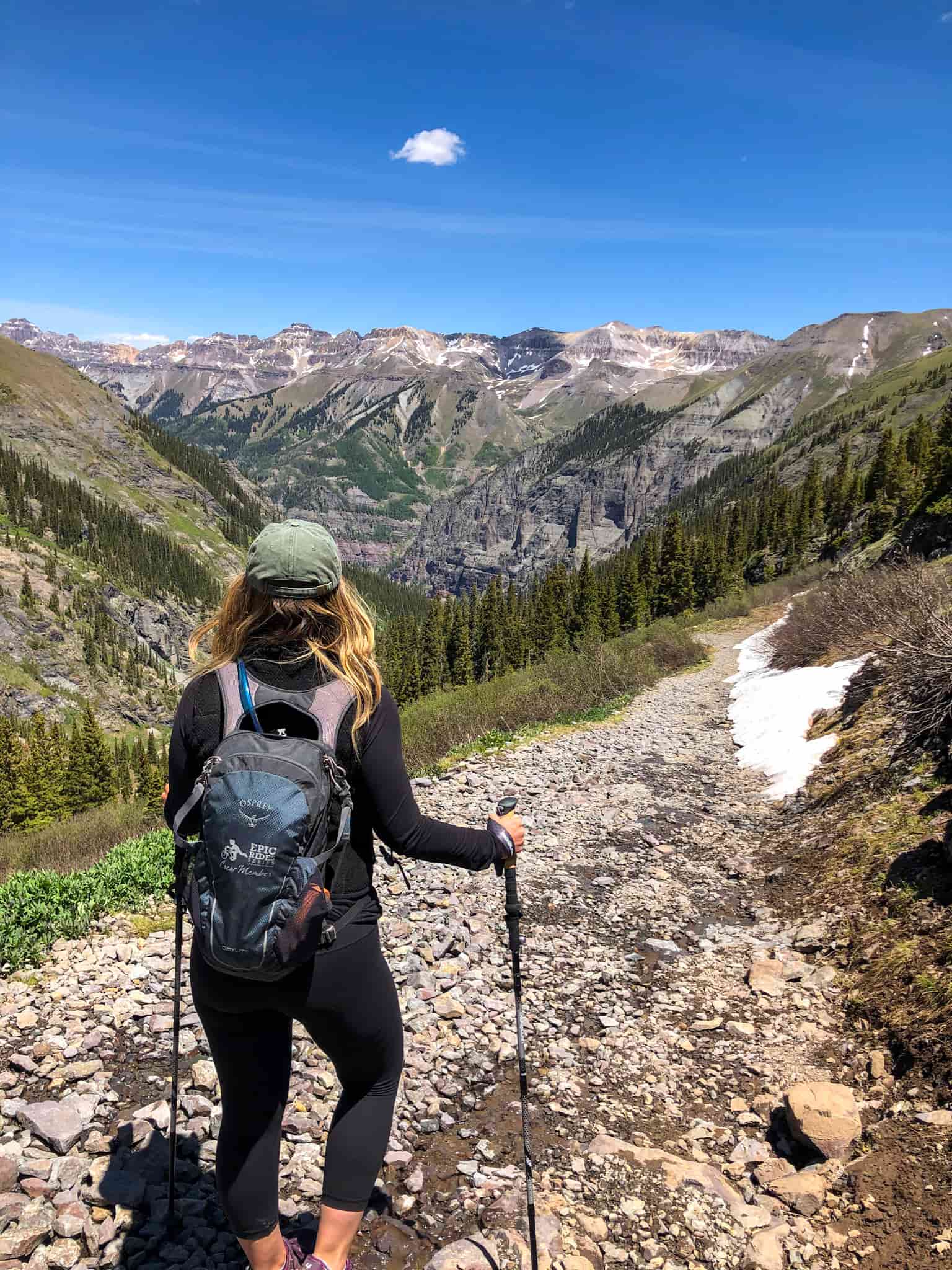 Woman wearing a backpack and carrying hiking poles stands on a rocky trail with mountains in the distance while hiking in Telluride, Colorado.