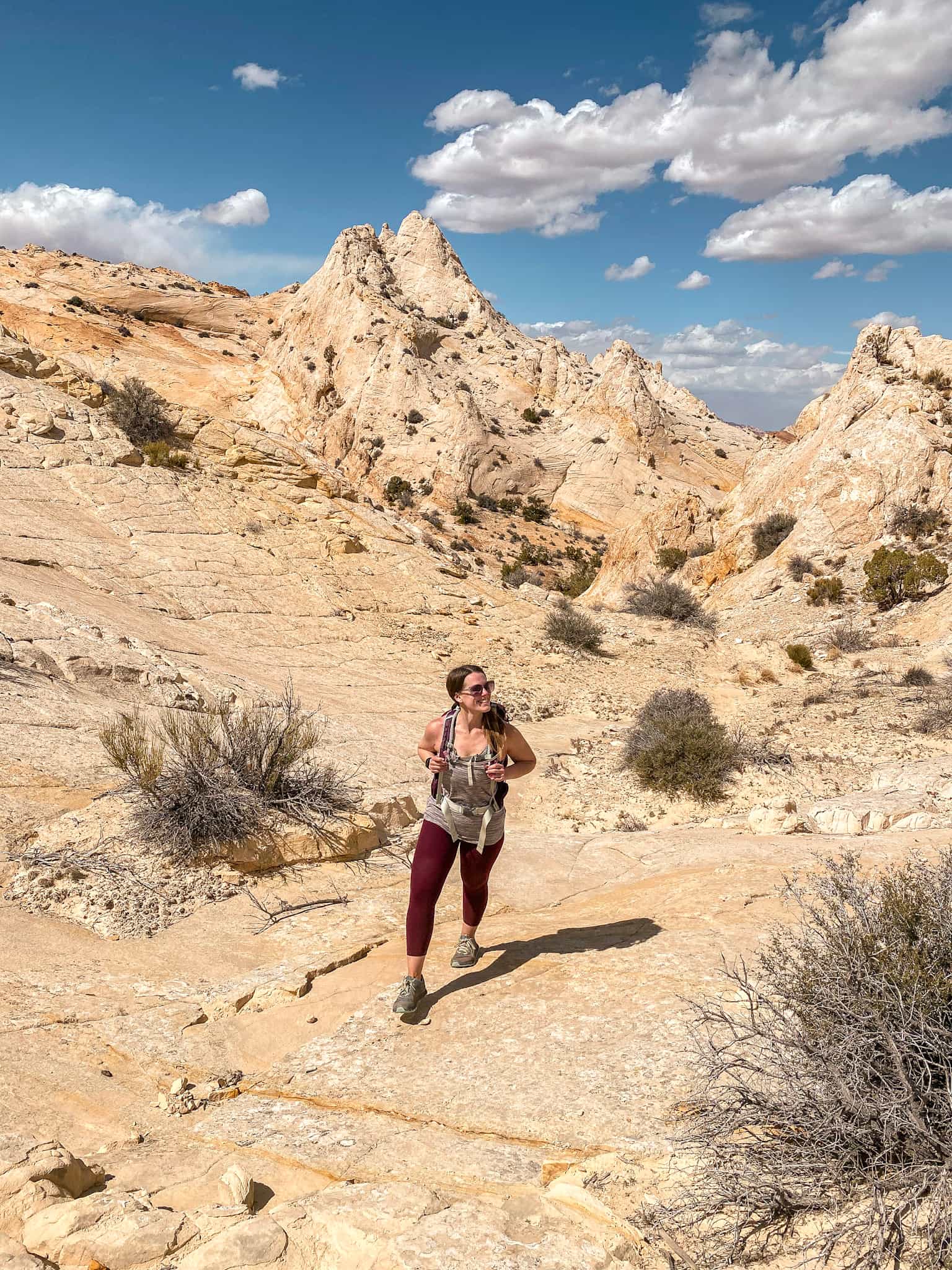 Female hiker wearing a backpack hiking across the desert with sandstone rock formations behind her in the Waterpocket Fold district of Capitol Reef National Park.